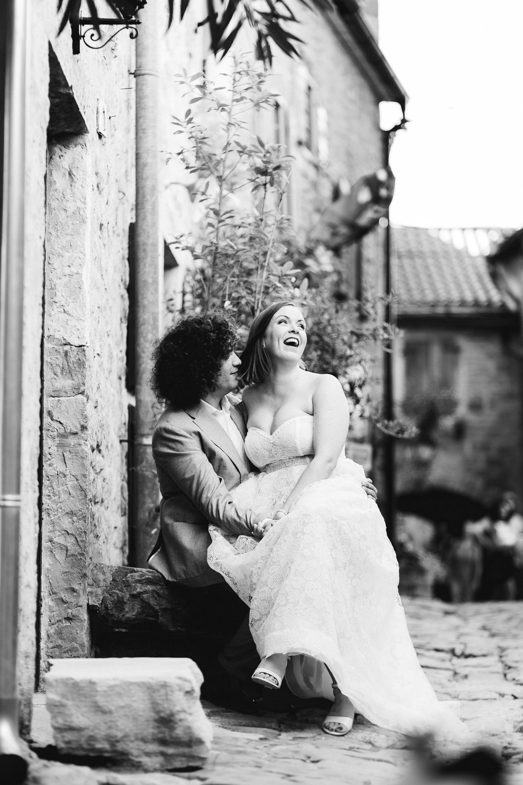 Mladenci u crno-bijeloj fotografiji tijekom vjenčanog sessiona u Grožnjanu, sjede na kamenoj klupi i smiju se u iskrenom trenutku ljubavi.